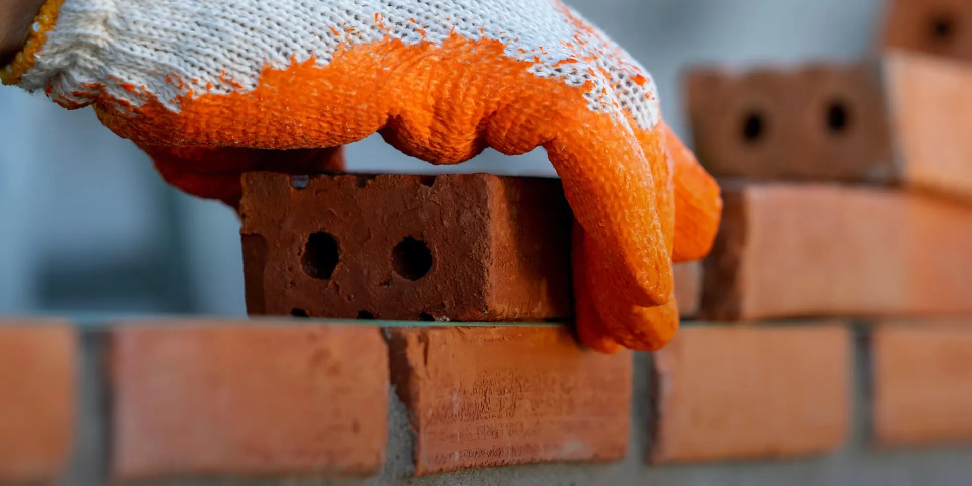 Statybininko ranka su oranžine pirštine deda plytą